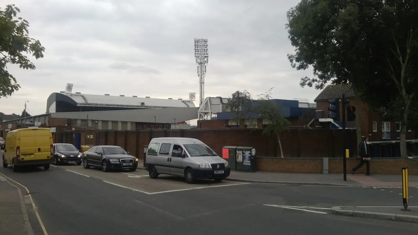 The curved roof of a stadium stand and a tall floodlight pylon rise behind a brick wall, with cars queuing on the road in front under a grey sky.