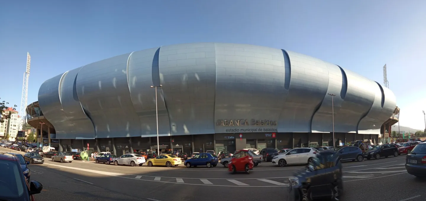 A wide view of the stadium's curved silver-clad facade bearing the name ABANCA Balaídos, with cars parked along the street in front under a clear blue sky.