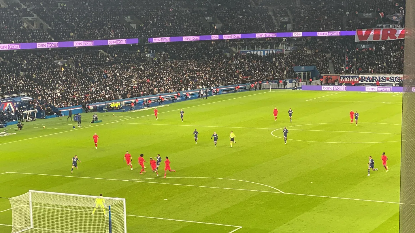 Elevated view of players in red and dark blue kits spread across a floodlit pitch in front of a packed stand, with a goal in the lower left corner.