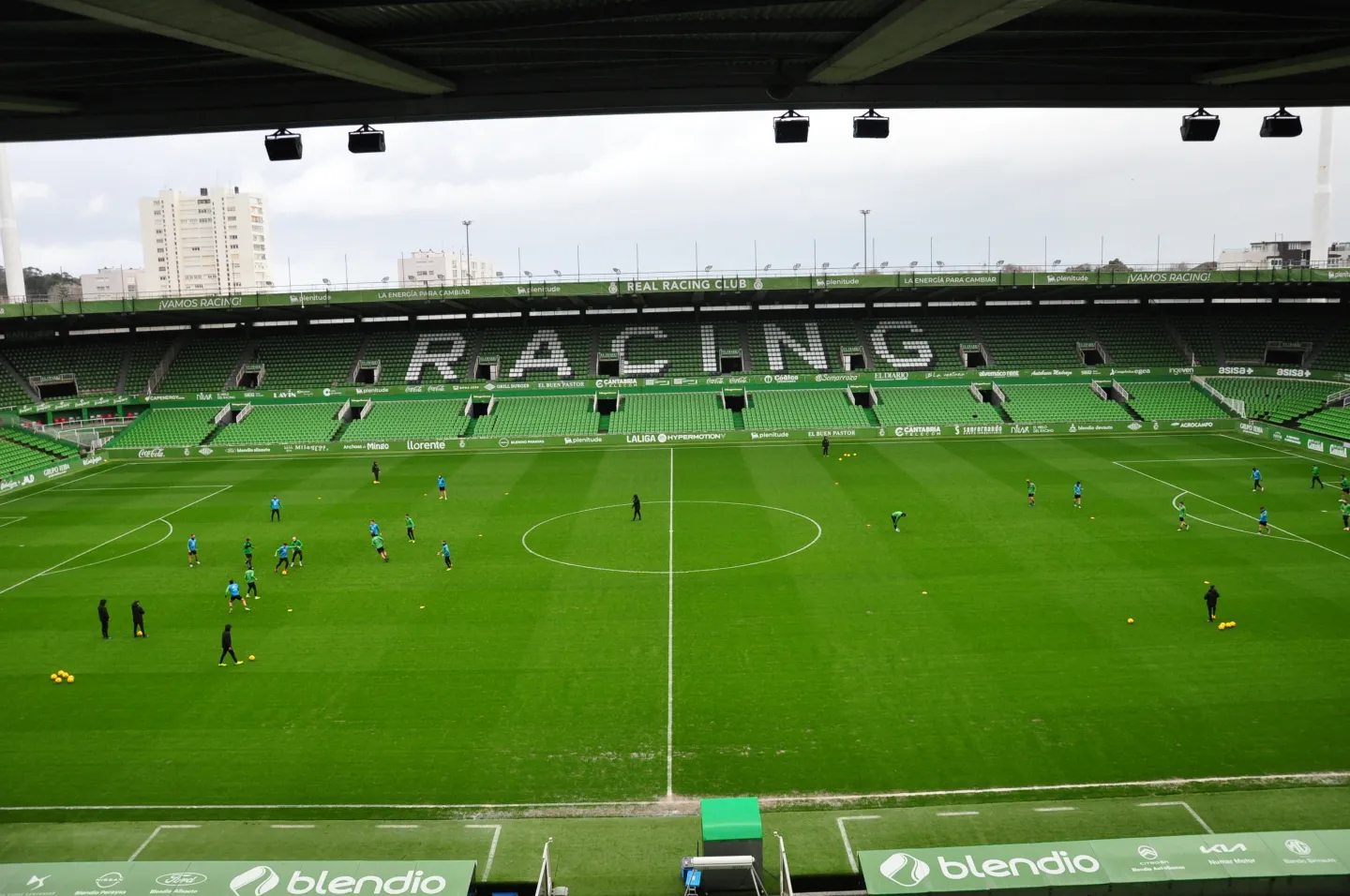A green football pitch with small groups of players in the foreground and green seating marked RACING across the opposite stand.