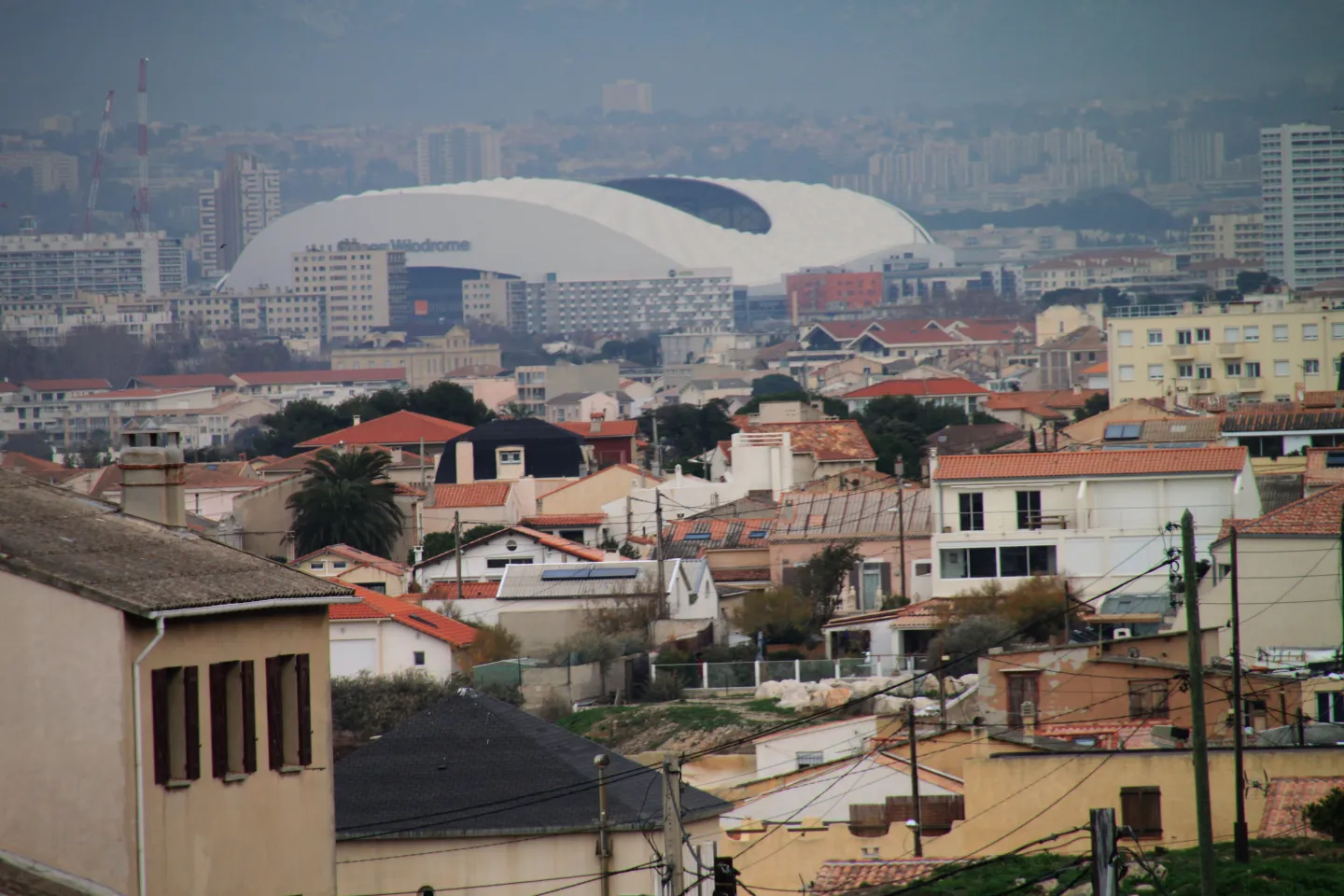 The stadium’s sweeping white roof rises behind residential buildings, with hills and a hazy sky in the distance.
